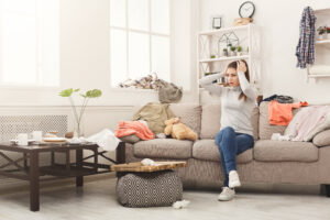 Desperate helpless woman sitting on sofa in messy living room. Young girl surrounded by many stack of clothes. Disorder and mess at home, copy space